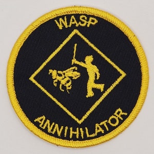 May include: A yellow and black embroidered patch with a diamond shape in the center. The diamond shape has a stick figure holding a stick and a wasp. The text "WASP ANNIHILATOR" is around the outside of the diamond shape.