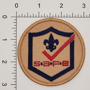 Puede incluir: Un parche redondo de color beige con una marca de verificaci&oacute;n azul y roja dentro de un escudo azul. El escudo tiene una flor de lis en el centro y la palabra "SAFE" en letras rojas debajo de la marca de verificaci&oacute;n.