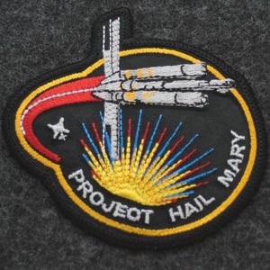 Puede incluir: Parche bordado con fondo negro, con un dise&ntilde;o de nave espacial y el texto "PROJECT HAIL MARY". El parche tiene un borde amarillo e incluye detalles en rojo, azul y amarillo, con una estrella y un sol estilizado.
