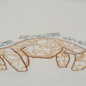 May include: Embroidered design featuring a stylized crab in shades of brown, with the word "AMAZE" arched above it in blue thread. The crab's body has intricate line details, set against a white background.
