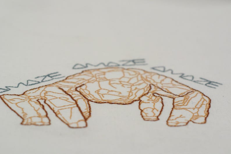 May include: Embroidered design featuring a stylized crab in shades of brown, with the word "AMAZE" arched above it in blue thread. The crab's body has intricate line details, set against a white background.