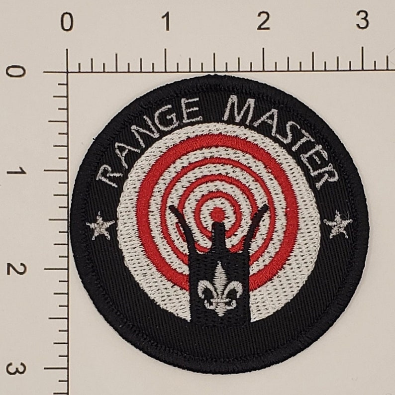 May include: Black and white embroidered patch with a red and white target in the centre. The text "RANGE MASTER" is written around the target in a white circle.