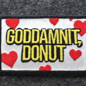 May include: Embroidered patch with the text "GODDAMNIT, DONUT" in yellow and black, surrounded by red heart shapes. The patch has a white background and a black border. The patch is rectangular.