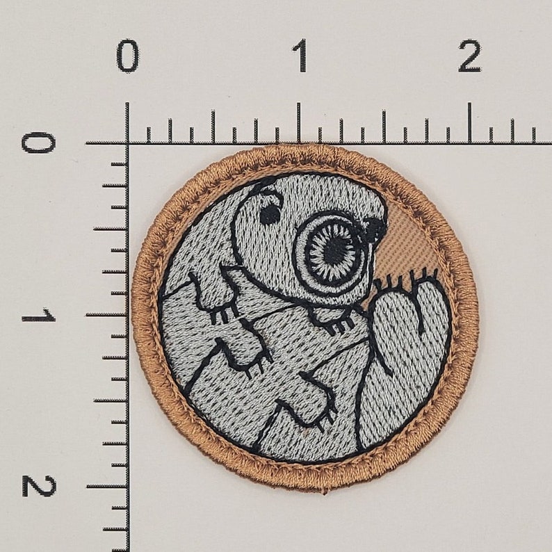K&ouml;nnte beinhalten: Gestickter Aufn&auml;her mit einem stilisierten B&auml;rtierchen, auch Tardigrade genannt, in Grau- und Schwarzt&ouml;nen. Der runde Aufn&auml;her hat einen braunen Rand und einen braunen Hintergrund. Der Aufn&auml;her hat einen Durchmesser von ca. 2,5 cm.