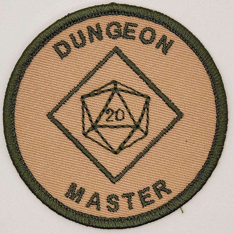 K&ouml;nnte beinhalten: Ein beigefarbener Stoff-Patch mit gr&uuml;nem gesticktem Text, der "DUNGEON MASTER" lautet, und einem gr&uuml;nen gestickten 20-seitigen W&uuml;rfel in der Mitte.