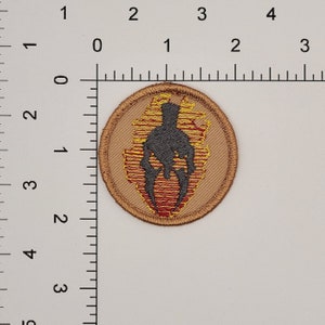 May include: A round, tan fabric patch with a black silhouette of a warrior in a helmet, surrounded by yellow and red flames.