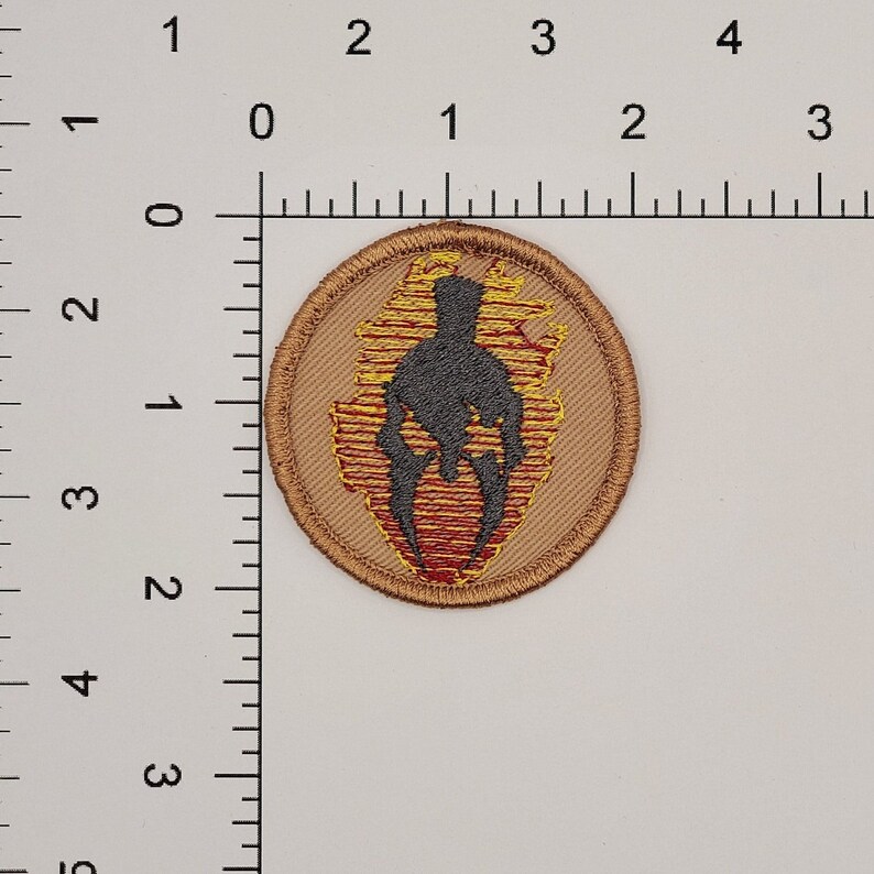 May include: A round, tan fabric patch with a black silhouette of a warrior in a helmet, surrounded by yellow and red flames.