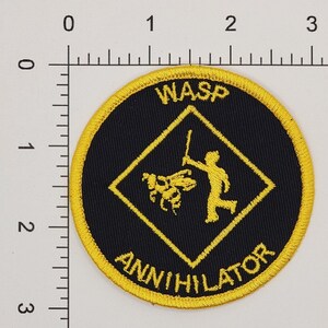 May include: A black and yellow embroidered patch with a yellow diamond shape in the center. The diamond shape shows a stick figure holding a fly swatter and a bee. The text "WASP ANNIHILATOR" is around the outside of the patch.