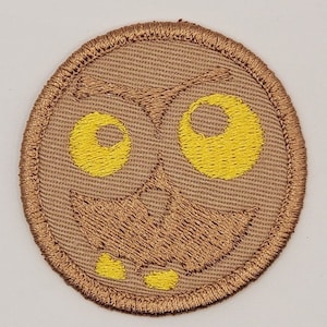 May include: A circular embroidered patch featuring a stylized owl face. The design includes large yellow eyes, a brown beak, and a brown outline. The patch has a tan background and a brown border.