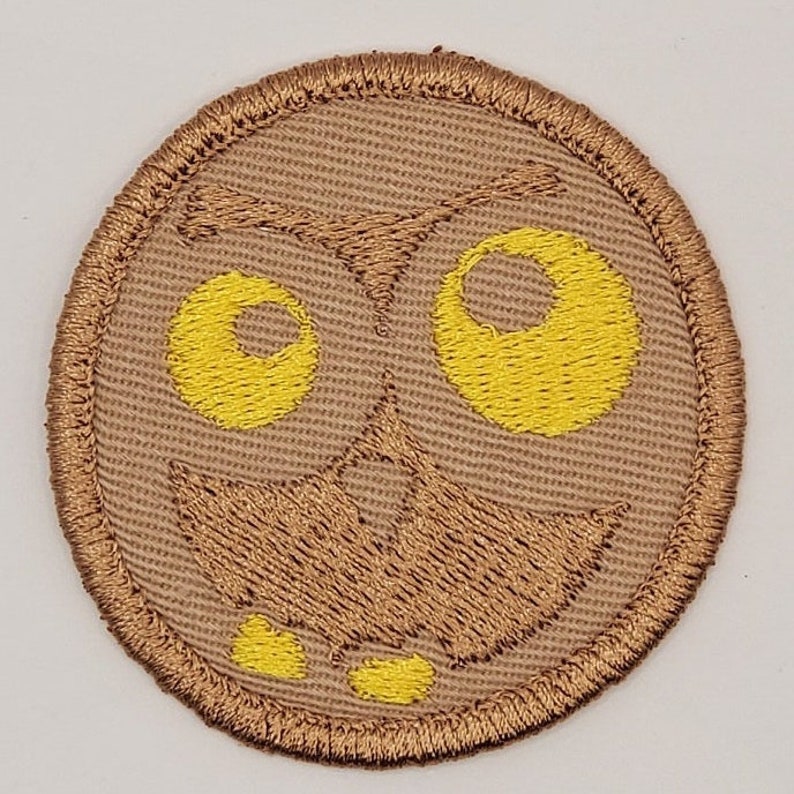 May include: A circular embroidered patch featuring a stylized owl face. The design includes large yellow eyes, a brown beak, and a brown outline. The patch has a tan background and a brown border.