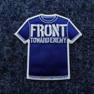 May include: A navy blue patch in the shape of a t-shirt with white stitching. The patch features the words "FRONT TOWARD ENEMY" in white block letters.