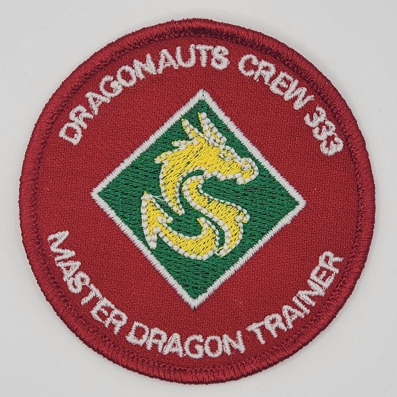 Peut inclure: &Eacute;cusson brod&eacute; rouge avec un contour blanc. L'&eacute;cusson pr&eacute;sente un diamant vert avec un dragon jaune &agrave; l'int&eacute;rieur. Le texte "Dragonaunts Crew 383 Master Dragon Trainer" est &eacute;crit en blanc autour du bord de l'&eacute;cusson.