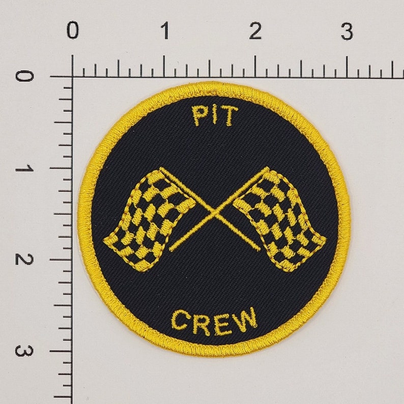 May include: A black and yellow circular patch with the words "PIT CREW" and two crossed checkered flags.