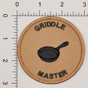 May include: A tan and green embroidered patch with the text "GRILL MASTER" and a black and white illustration of a frying pan.