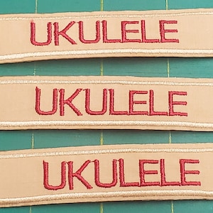 May include: Three rectangular patches with the word "UKULELE" embroidered in red thread on a tan background. Each patch has a white embroidered border. The patches are arranged on a green cutting mat.