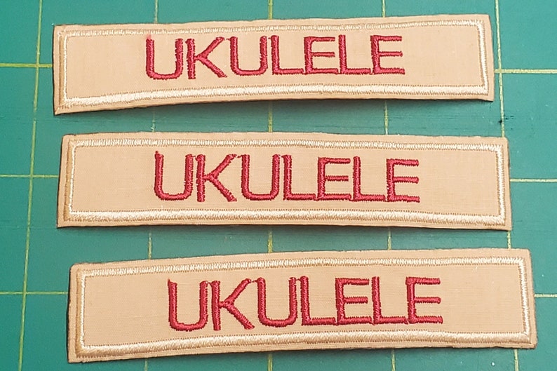 May include: Three rectangular patches with the word "UKULELE" embroidered in red thread on a tan background. Each patch has a white embroidered border. The patches are arranged on a green cutting mat.