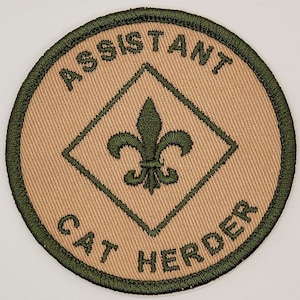 May include: A tan and green embroidered patch with the text "Assistant Cat Herder" and a green fleur-de-lis in a diamond shape.