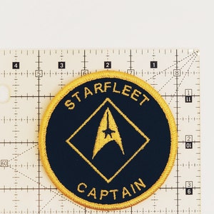 May include: A round, black and gold embroidered patch with the text "STARFLEET" and "CAPTAIN" in a yellow circle. A gold starfleet insignia is in the center of the patch.