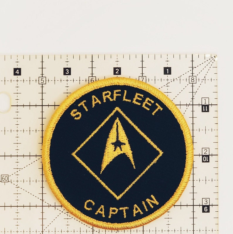 May include: A round, black and gold embroidered patch with the text "STARFLEET" and "CAPTAIN" in a yellow circle. A gold starfleet insignia is in the center of the patch.