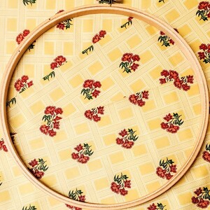 Pu&ograve; includere: Un telaio da ricamo in legno con finitura in legno naturale. Il telaio &egrave; appoggiato su un tessuto giallo con un motivo floreale.
