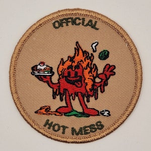 May include: A tan embroidered patch with a cartoon character in flames holding a slice of cake. The text "OFFICIAL HOT MESS" is embroidered around the character.