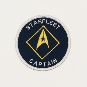 May include: A round, navy blue embroidered patch with a white border. The patch features a yellow diamond with a yellow starfleet insignia inside. The text "STARFLEET" is above the diamond and "CAPTAIN" is below.