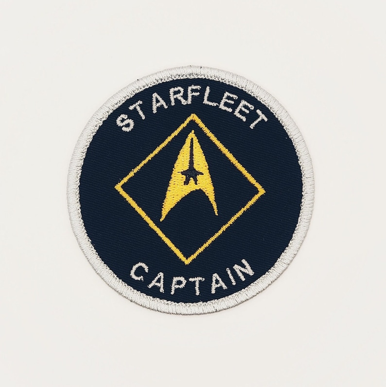 May include: A round, navy blue embroidered patch with a white border. The patch features a yellow diamond with a yellow starfleet insignia inside. The text "STARFLEET" is above the diamond and "CAPTAIN" is below.