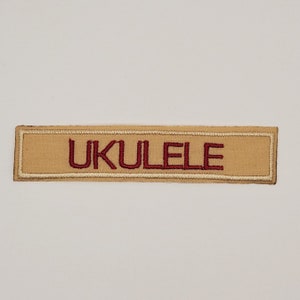 May include: A rectangular patch with the word "UKULELE" embroidered in burgundy thread on a tan background. The patch has a tan border and is likely for a musical instrument.