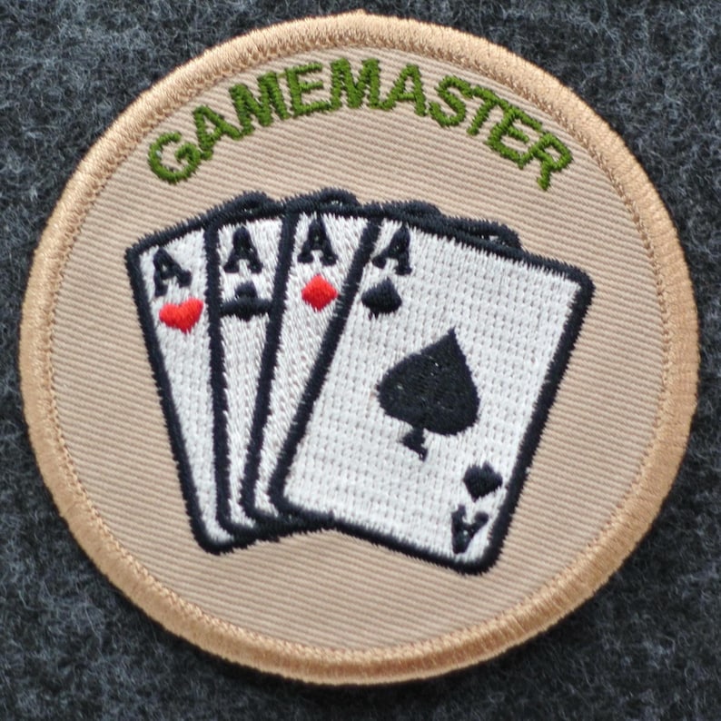 May include: A circular patch with the word "GAMEMASTER" embroidered in green above four playing cards. The cards are white with black details, featuring three aces of hearts and one ace of spades. The patch has a beige background and a beige border.