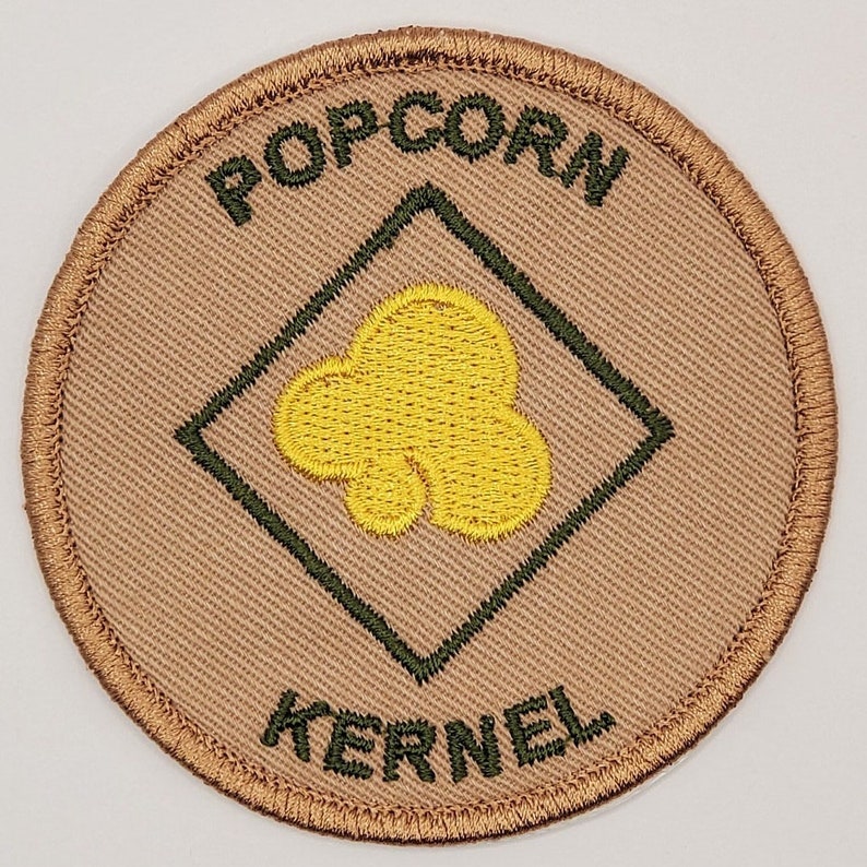 May include: A tan round patch with a green diamond in the center. The diamond has a yellow popcorn kernel inside. The words "POPCORN KERNEL" are embroidered around the outside of the patch.