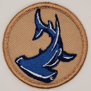May include: Round patch featuring a blue hammerhead shark design with white accents, centered on a tan background. The patch has a brown stitched border, suitable for sewing or ironing onto fabric. Ideal for marine life enthusiasts.