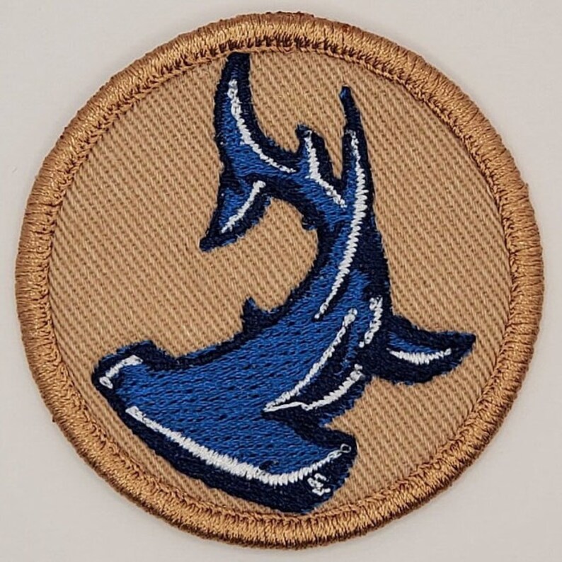 Op de afbeelding: Ronde patch met een blauw hamerhaai-ontwerp en witte accenten, op een beige achtergrond. De patch heeft een bruine gestikte rand. Geschikt om op stof te naaien of te strijken. Ideaal voor liefhebbers van het zeeleven.