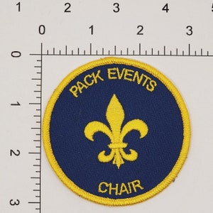 May include: A blue and yellow round patch with a fleur-de-lis in the center. The text "PACK EVENTS CHAIR" is written around the edge of the patch.