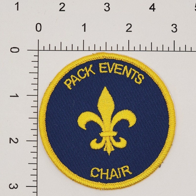 May include: A blue and yellow round patch with a fleur-de-lis in the center. The text "PACK EVENTS CHAIR" is written around the edge of the patch.