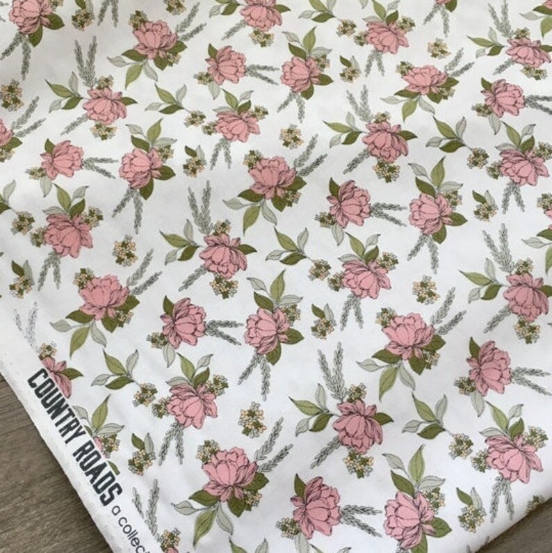 以下が含まれることがあります： 白い生地に繰り返しの花柄。デザインは、ピンクのシャクヤクのような花、緑の葉、麦の穂が特徴です。「COUNTRY ROADS a collection」の文字が生地の端に印刷されています。