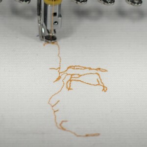 May include: Close-up of an embroidery machine in operation, stitching a design with orange thread onto white fabric. The needle is visible, creating the pattern. The image highlights the process of textile art.