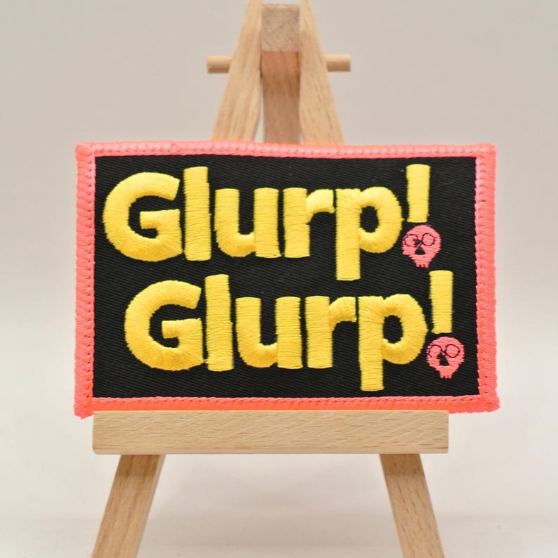 May include: Embroidered patch with the word "Glurp!" in yellow, repeated on a black background. The patch has a pink border and small skull graphics. The patch is displayed on a small wooden easel.