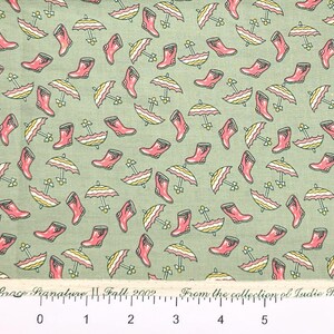 May include: A green fabric with a repeating pattern of pink rain boots and yellow and white umbrellas. The fabric has a ruler with inches marked on it. The text on the ruler reads "Aunt Grace Signature Fall 2002 From the collection of Ludie Rothermel."