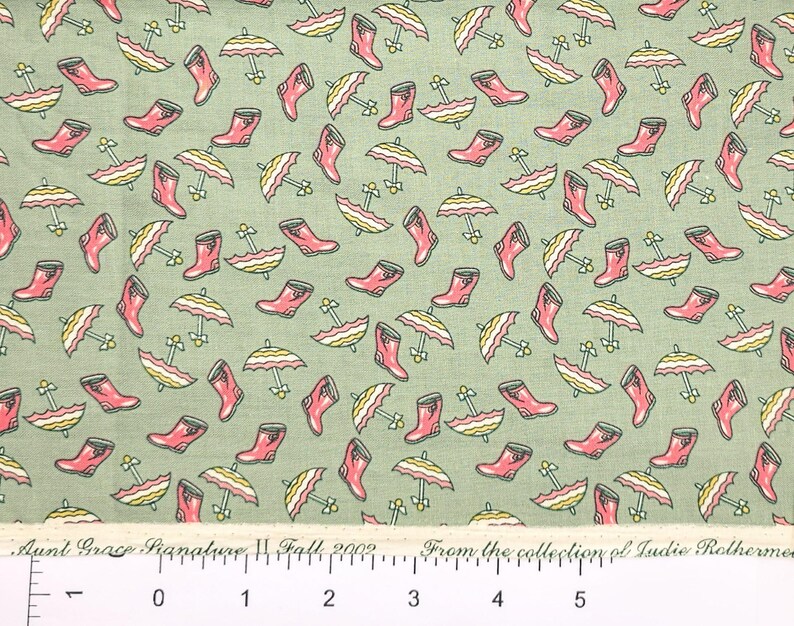 May include: A green fabric with a repeating pattern of pink rain boots and yellow and white umbrellas. The fabric has a ruler with inches marked on it. The text on the ruler reads "Aunt Grace Signature Fall 2002 From the collection of Ludie Rothermel."