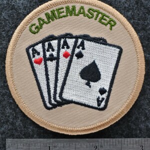 May include: A circular patch featuring the word "GAMEMASTER" in green above four playing cards, each showing an Ace. The cards have black and red suit symbols. The patch has a beige background and a beige border.