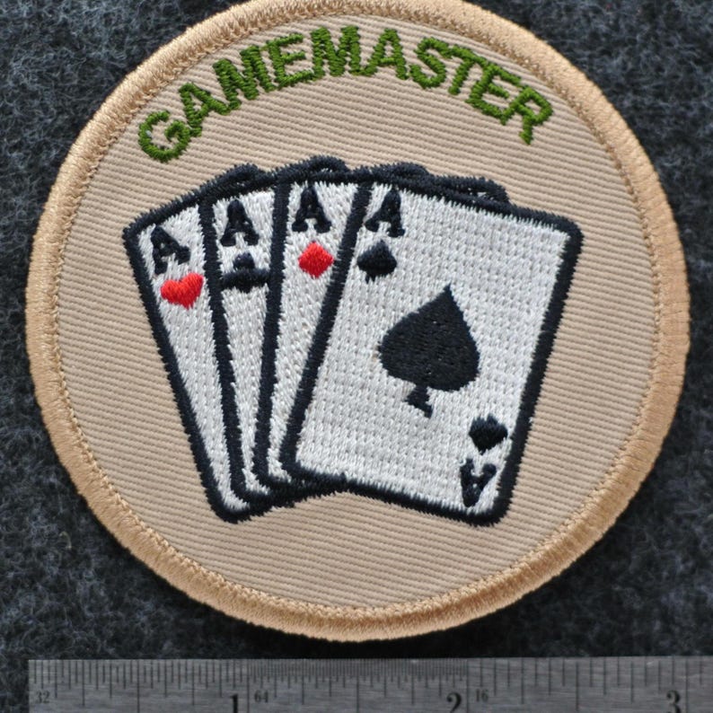 May include: A circular patch featuring the word "GAMEMASTER" in green above four playing cards, each showing an Ace. The cards have black and red suit symbols. The patch has a beige background and a beige border.