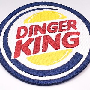 K&ouml;nnte beinhalten: Ein wei&szlig;er, blauer und roter gestickter Aufn&auml;her mit dem Text "Dinger King" in einer stilisierten Schriftart. Der Aufn&auml;her zeigt einen gelben und wei&szlig;en Kreis in der Mitte.