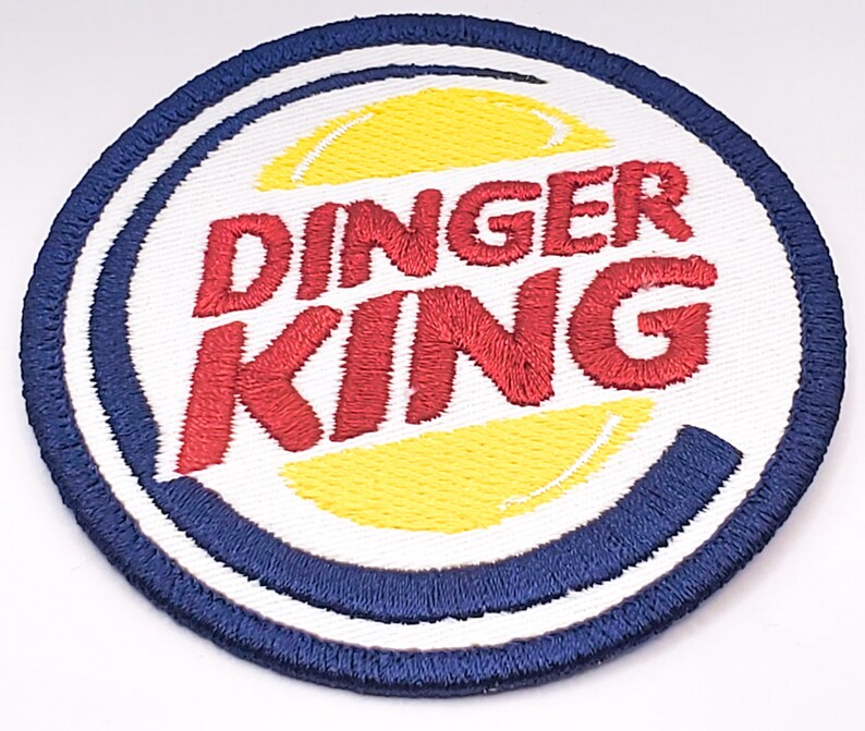 K&ouml;nnte beinhalten: Ein wei&szlig;er, blauer und roter gestickter Aufn&auml;her mit dem Text "Dinger King" in einer stilisierten Schriftart. Der Aufn&auml;her zeigt einen gelben und wei&szlig;en Kreis in der Mitte.