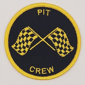 May include: A round, yellow embroidered patch with black fabric in the center. The patch has the words "PIT CREW" in yellow lettering and two crossed checkered flags in yellow.