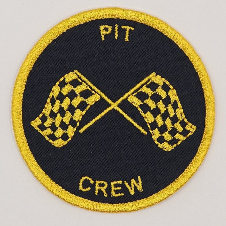May include: A round, yellow embroidered patch with black fabric in the center. The patch has the words "PIT CREW" in yellow lettering and two crossed checkered flags in yellow.