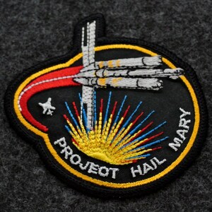 Puede incluir: Parche bordado con fondo negro, con una nave espacial estilizada y el texto "PROJECT HAIL MARY". El dise&ntilde;o incluye una combinaci&oacute;n de colores amarillo, rojo, azul y blanco, con una estrella y un motivo de sol naciente.