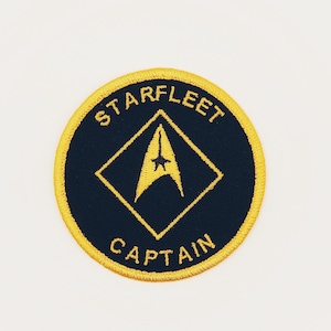 May include: A round, black embroidered patch with a yellow border. The patch features a yellow diamond shape with a yellow starfleet insignia inside. The text "STARFLEET" is above the diamond and "CAPTAIN" is below.