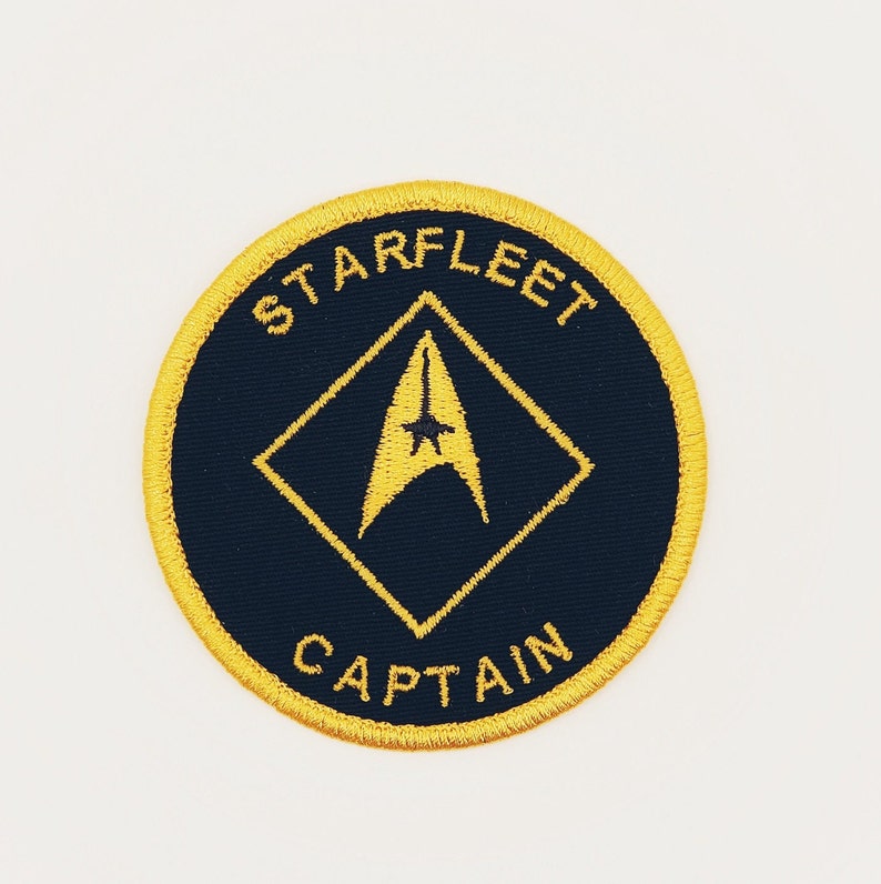 May include: A round, black embroidered patch with a yellow border. The patch features a yellow diamond shape with a yellow starfleet insignia inside. The text "STARFLEET" is above the diamond and "CAPTAIN" is below.