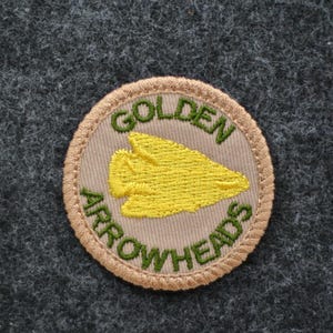 Pu&ograve; includere: Toppa circolare con le parole "GOLDEN ARROWHEADS" ricamate in verde su sfondo beige. Una punta di freccia gialla &egrave; al centro. La toppa ha un bordo beige ed &egrave; posta su un tessuto grigio strutturato.