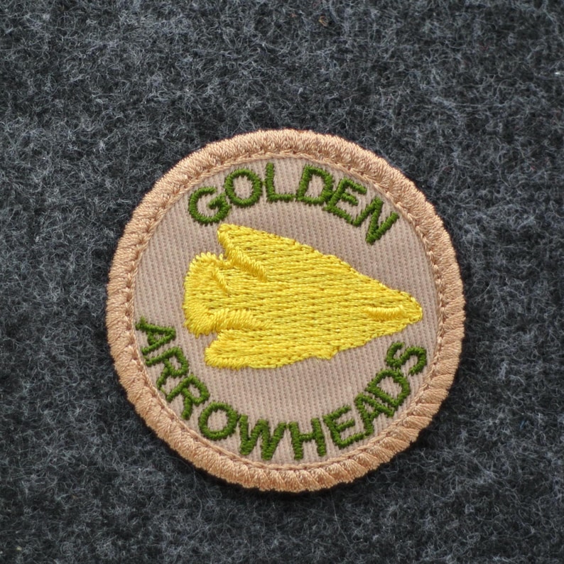 Pu&ograve; includere: Toppa circolare con le parole "GOLDEN ARROWHEADS" ricamate in verde su sfondo beige. Una punta di freccia gialla &egrave; al centro. La toppa ha un bordo beige ed &egrave; posta su un tessuto grigio strutturato.